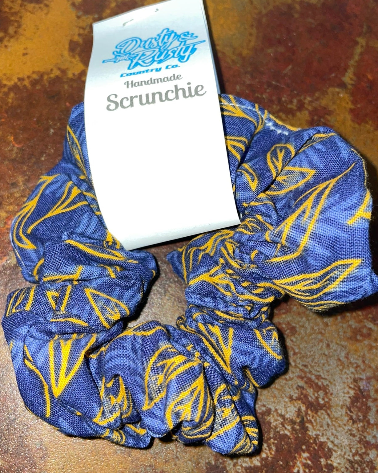 A blue and yellow patterned fabric scrunchie with a white paper tag that reads 'Handmade Scrunchie' on a textured brown surface.