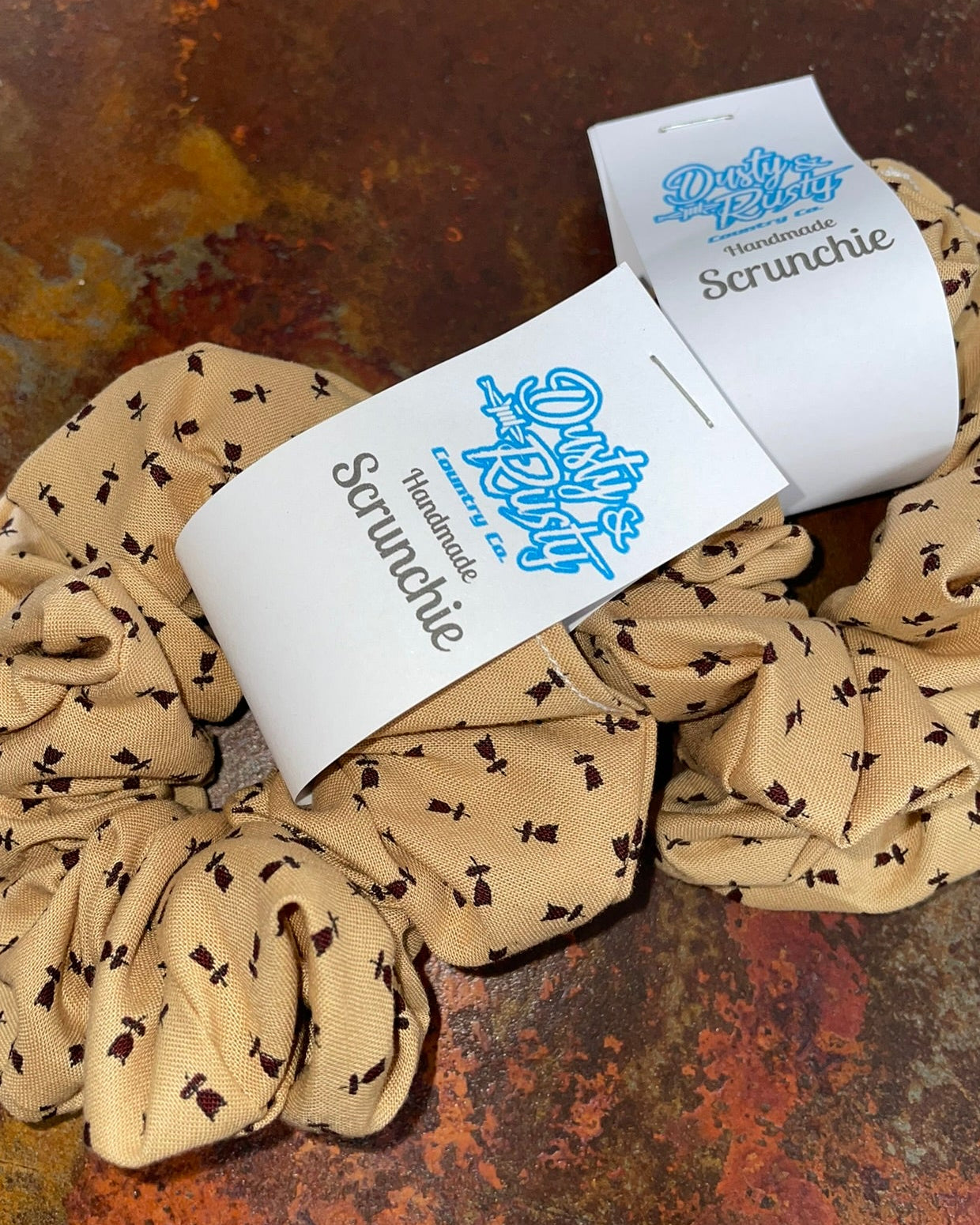 A beige scrunchie with a small black dog print pattern, featuring a white paper tag printed with the brand name 'Dusty Paws' and the word 'Scrunchie'. The scrunchie is displayed on a textured, rusty brown surface.