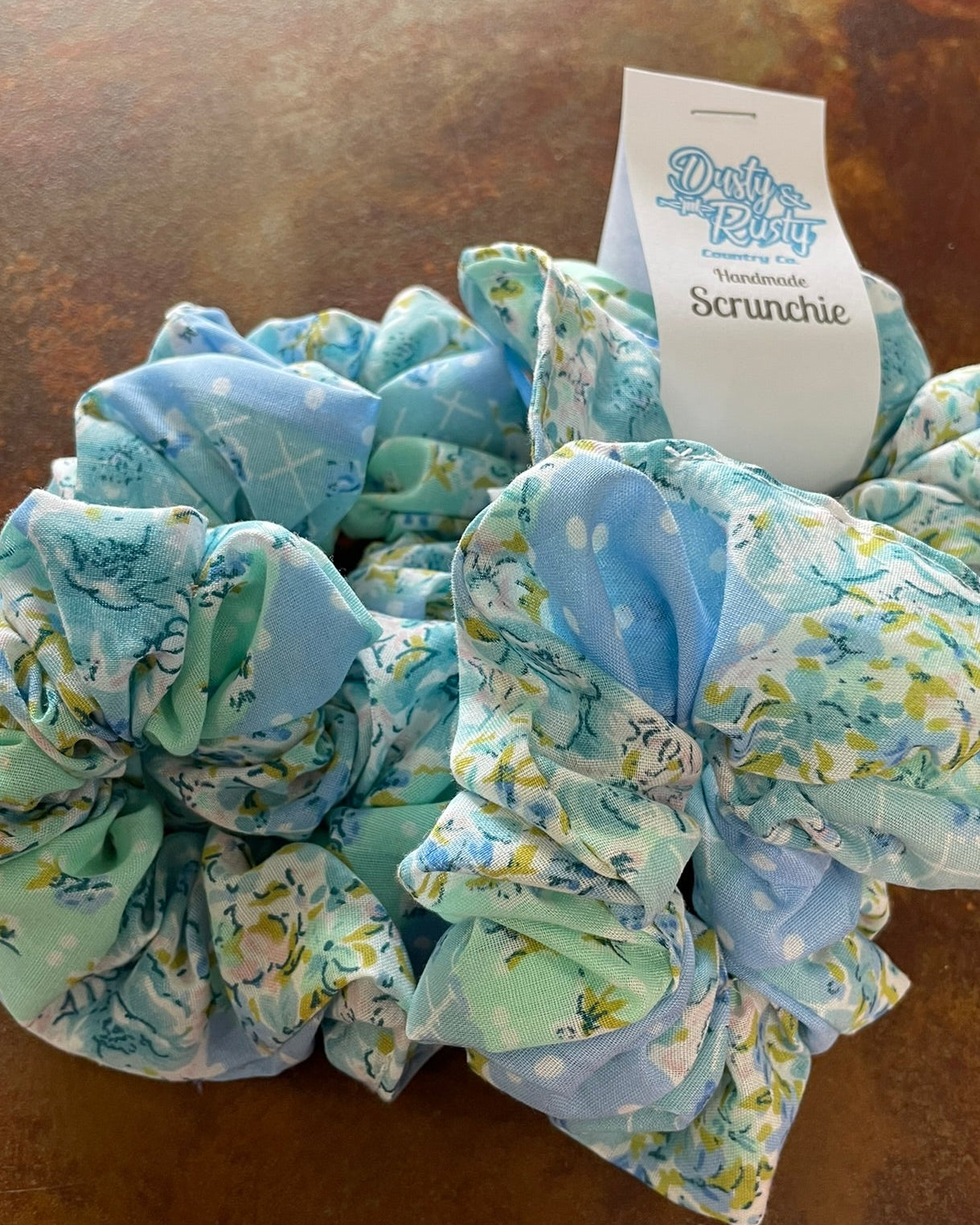 Three fabric hair scrunchies in light blue and mint green floral print with a white paper tag that reads 'Dusty Petal Scrunchie' on a brown textured surface.