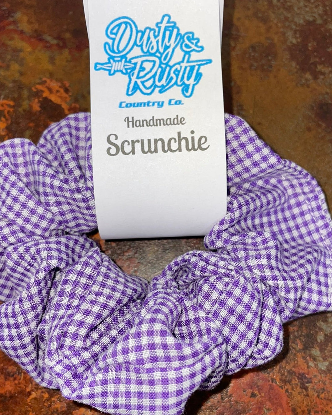 A purple and white gingham fabric scrunchie with a white paper tag that reads 'Dirty Riff Country Co. Handmade Scrunchie' on a textured rusted metal surface.