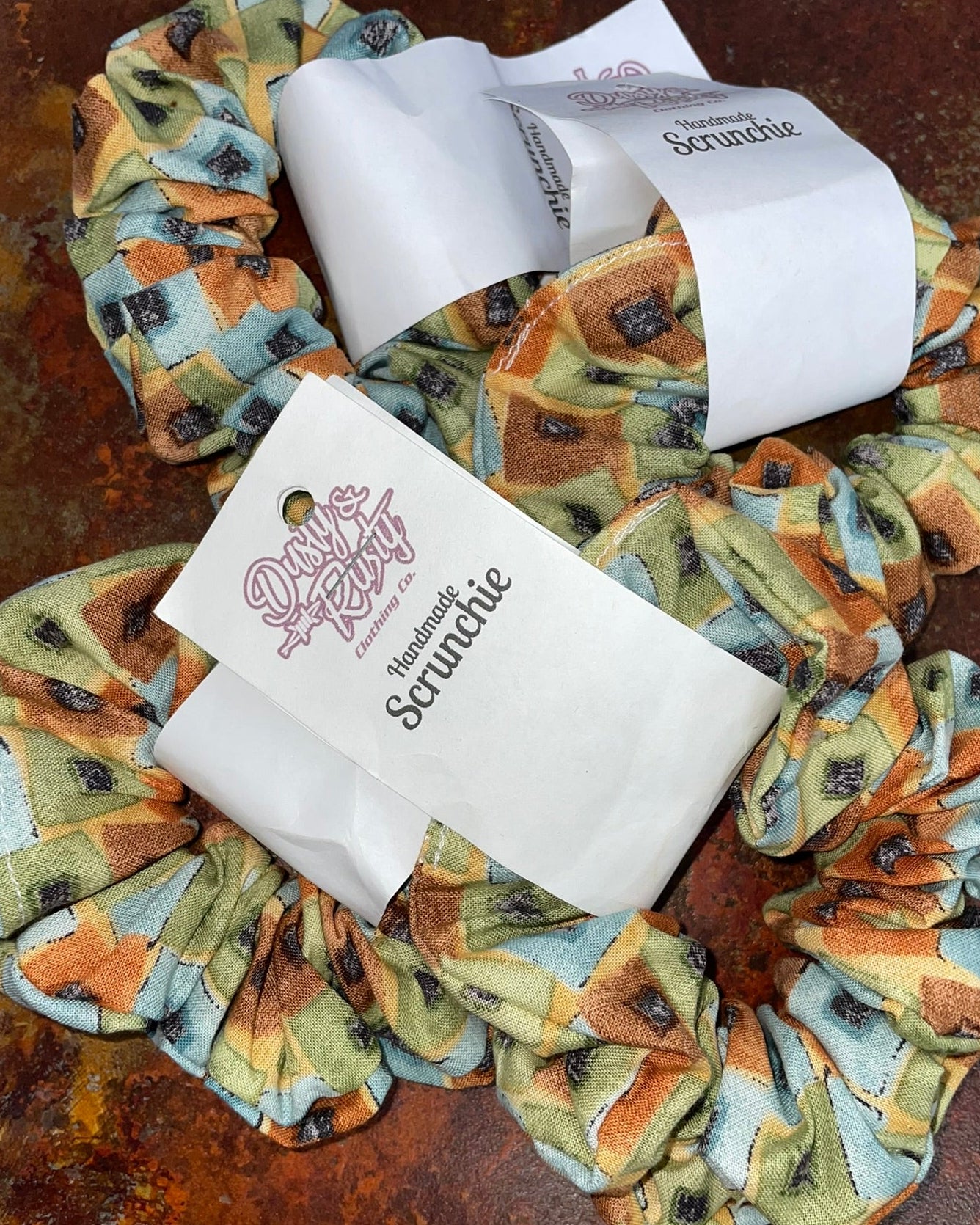 Two fabric scrunchies with a vintage-style floral and botanical print in muted green, orange, and beige tones, each wrapped with a white paper band labeled 'Dusty Rose Scrunchie'.