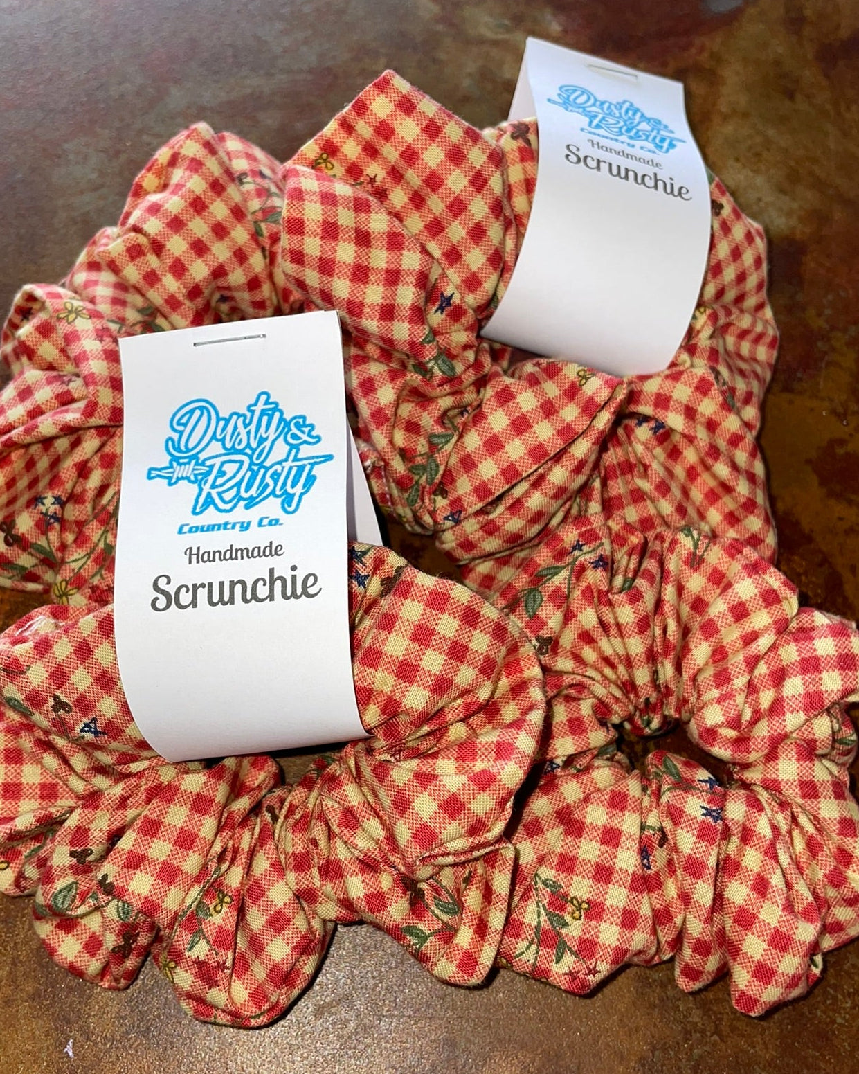 Three red and white gingham scrunchies with white paper tags on a dark brown textured surface. The tags read 'Dusty Dog' and 'Handmade Scrunchie'.