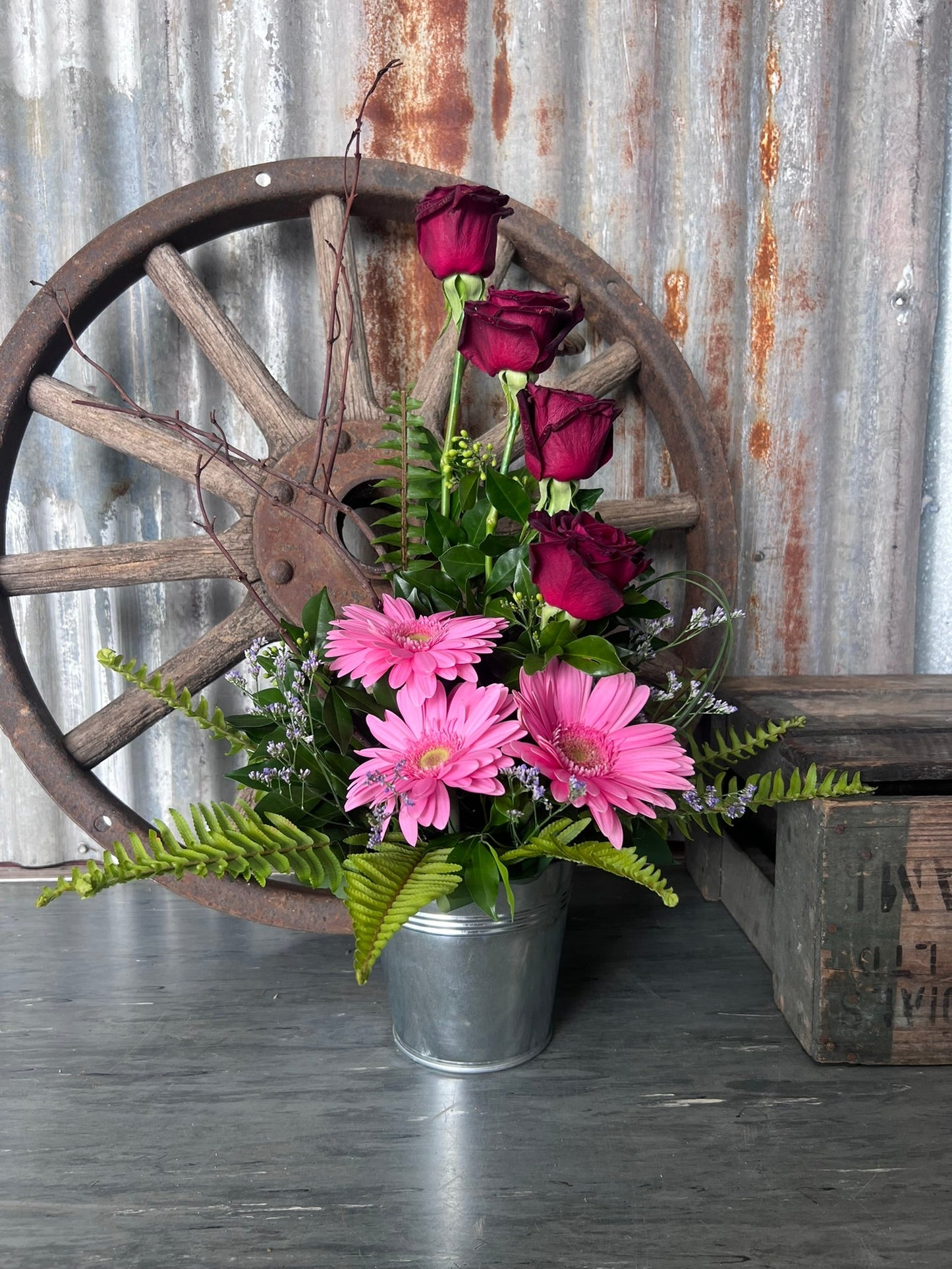 Floral arrangement in a metal container with a rustic wooden wagon wheel and wooden crate in the background.
