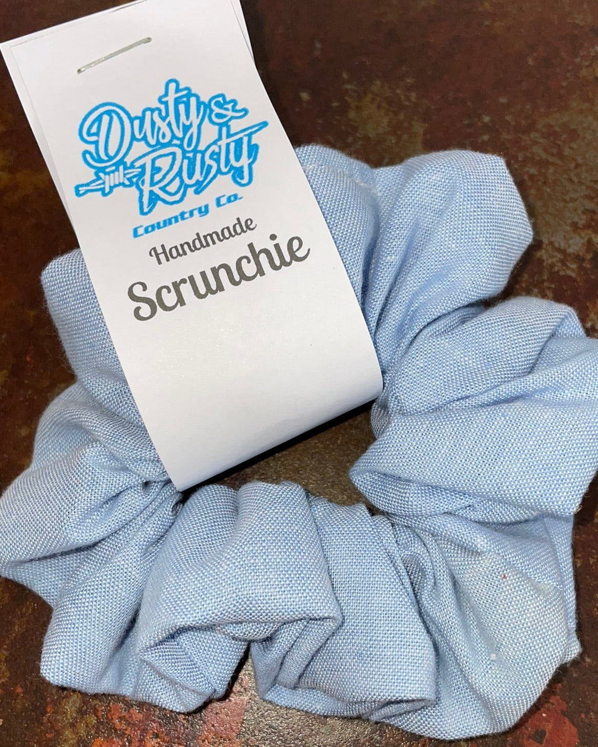 A light blue fabric scrunchie with a white paper tag that reads 'Dusty Rusty Country Co Handmade Scrunchie,' placed on a rusted metal surface.