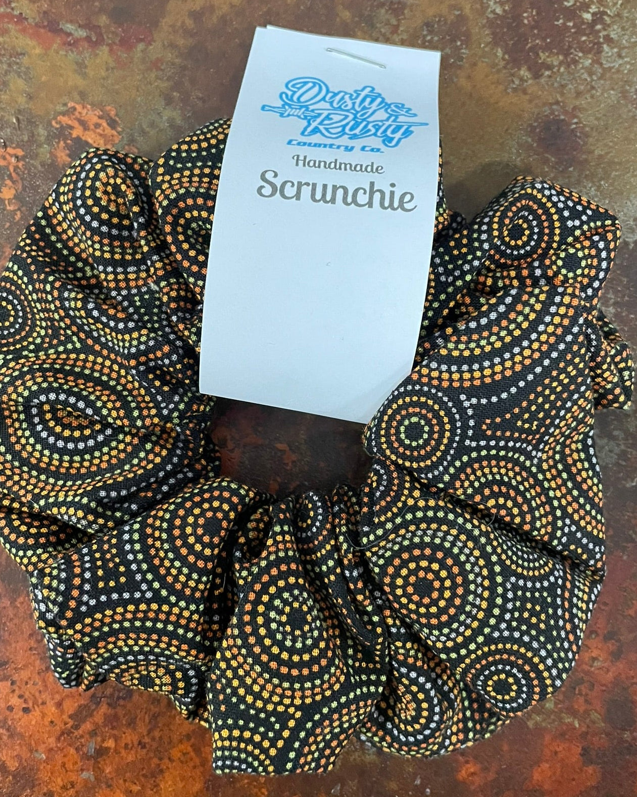 A handmade fabric scrunchie with a black background and gold and white dot pattern, featuring a white paper tag that reads 'Dusty Betty Handmade Scrunchie'.