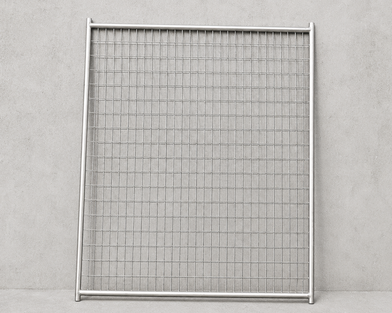 A square metal mesh fence panel with a silver aluminum frame, standing upright against a textured gray concrete wall.