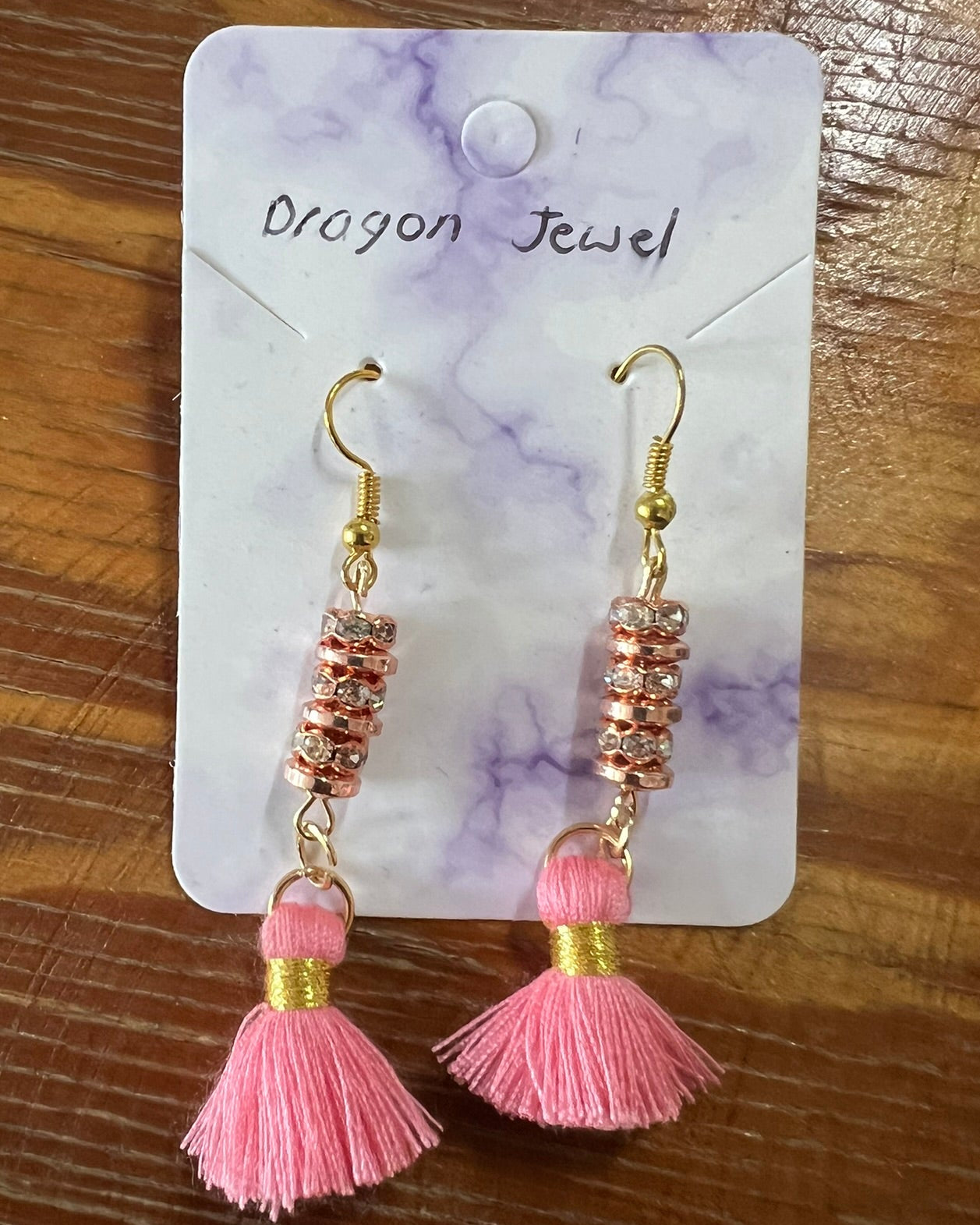 Pink tassel earrings on a 'Dragon Jewel' branded card against a wooden background