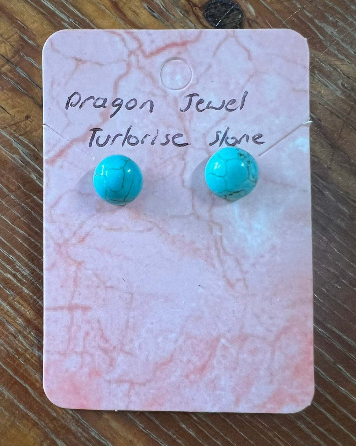 Turquoise stone earrings on a pink marble card with 'Dragon Jewel' text, placed on a wooden surface.