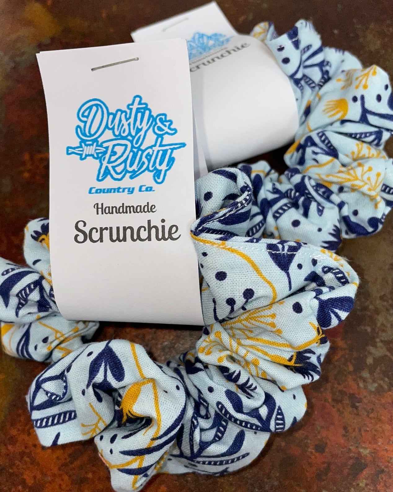 A handmade fabric scrunchie with a white background and navy blue nautical print, featuring a white paper tag with the brand 'Dusty's Rust Country Co.' printed in blue and black.