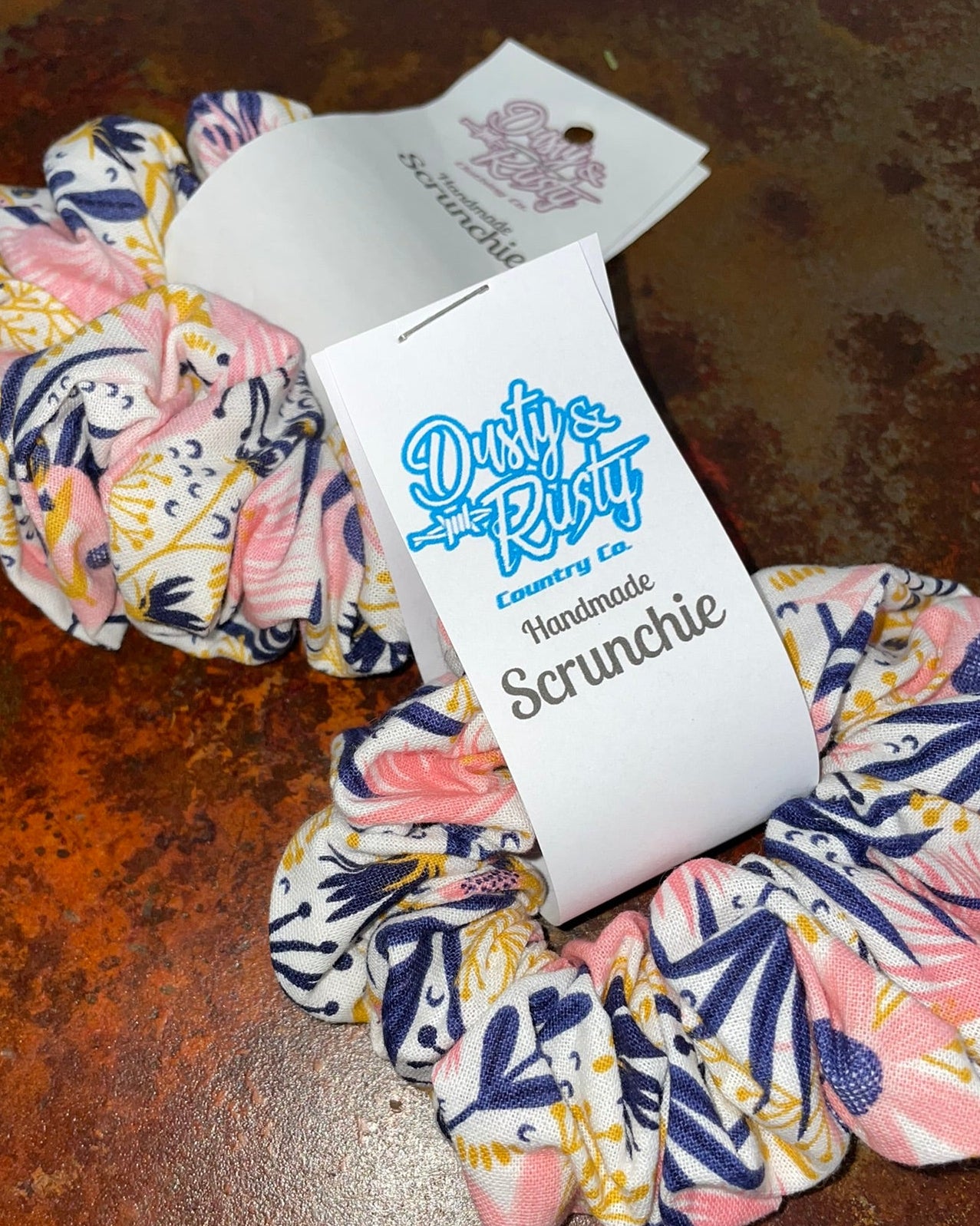 Two fabric scrunchies with a pink, navy, and yellow botanical pattern, each attached to a white paper tag. One tag reads 'Dirty Riffle Handmade Scrunchie' and the other shows the 'Sea Sandwich' brand logo.