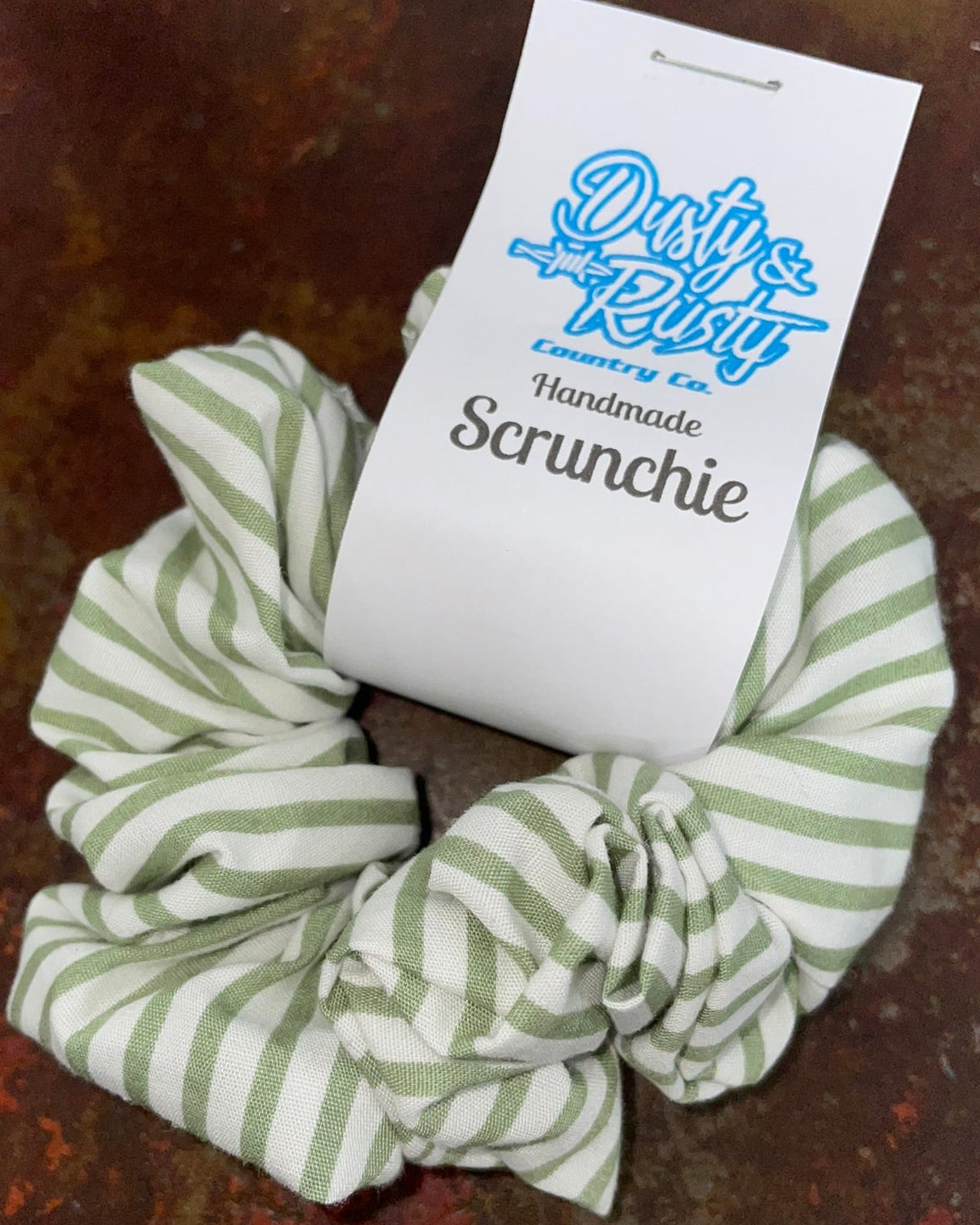 A fabric scrunchie with light green and white stripes, featuring a white paper tag attached that reads 'Dusty Rhy Handmade Scrunchie' in blue and black text, set against a rustic brown textured background.