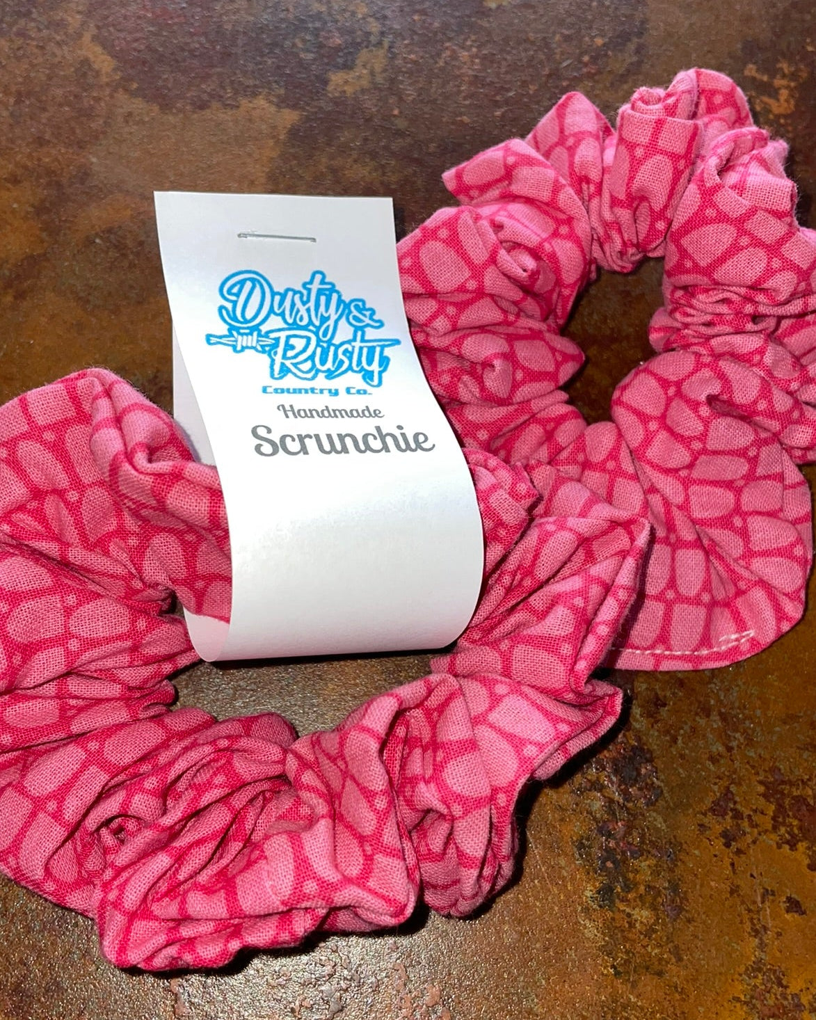 A pink scrunchie with a lighter pink abstract pattern, featuring a white paper tag with the brand name 'Dusty Rose Handmade Scrunchie' on a rustic brown background.