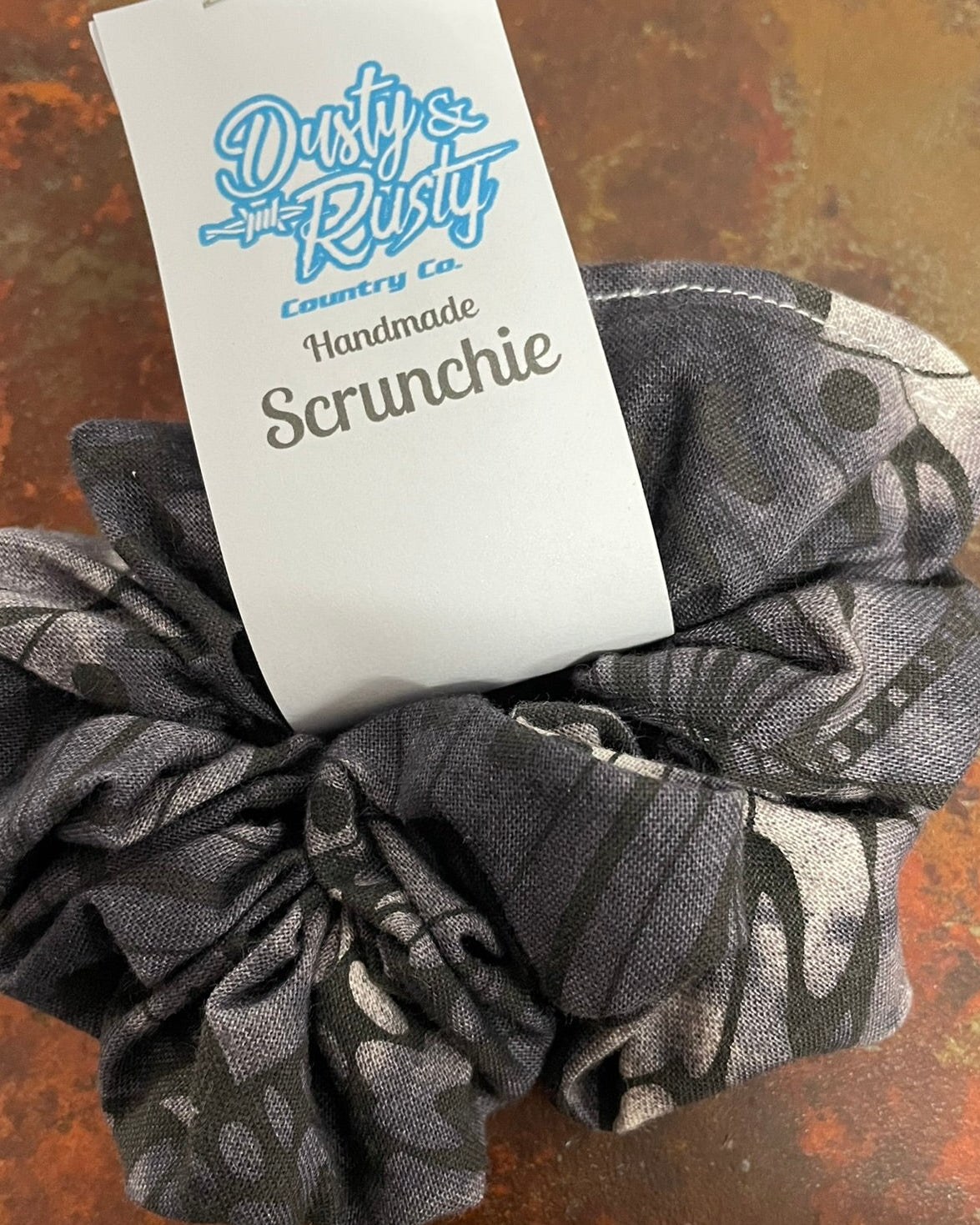 A dark gray scrunchie with a black and gray abstract pattern, featuring a white paper tag that reads 'Dusty Rusty Country Co Handmade Scrunchie' on a textured rusted metal surface.