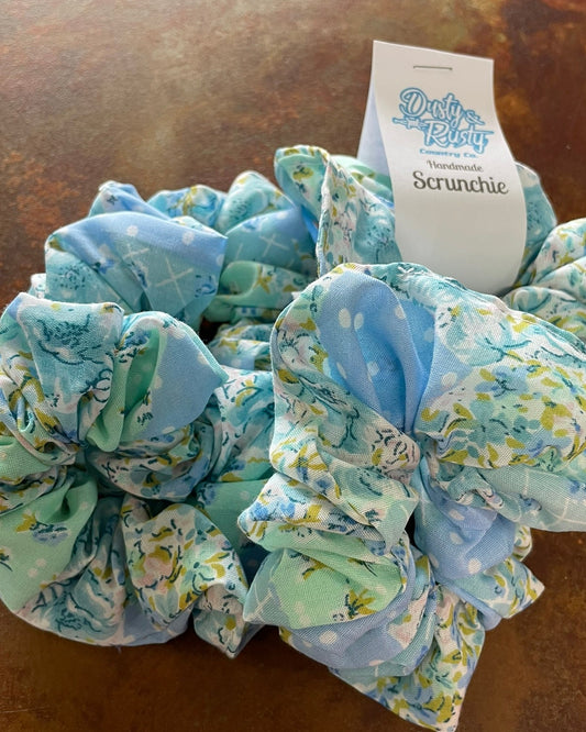 Three fabric hair scrunchies in light blue and mint green floral print with a white paper tag that reads 'Dusty Petal Scrunchie' on a brown textured surface.