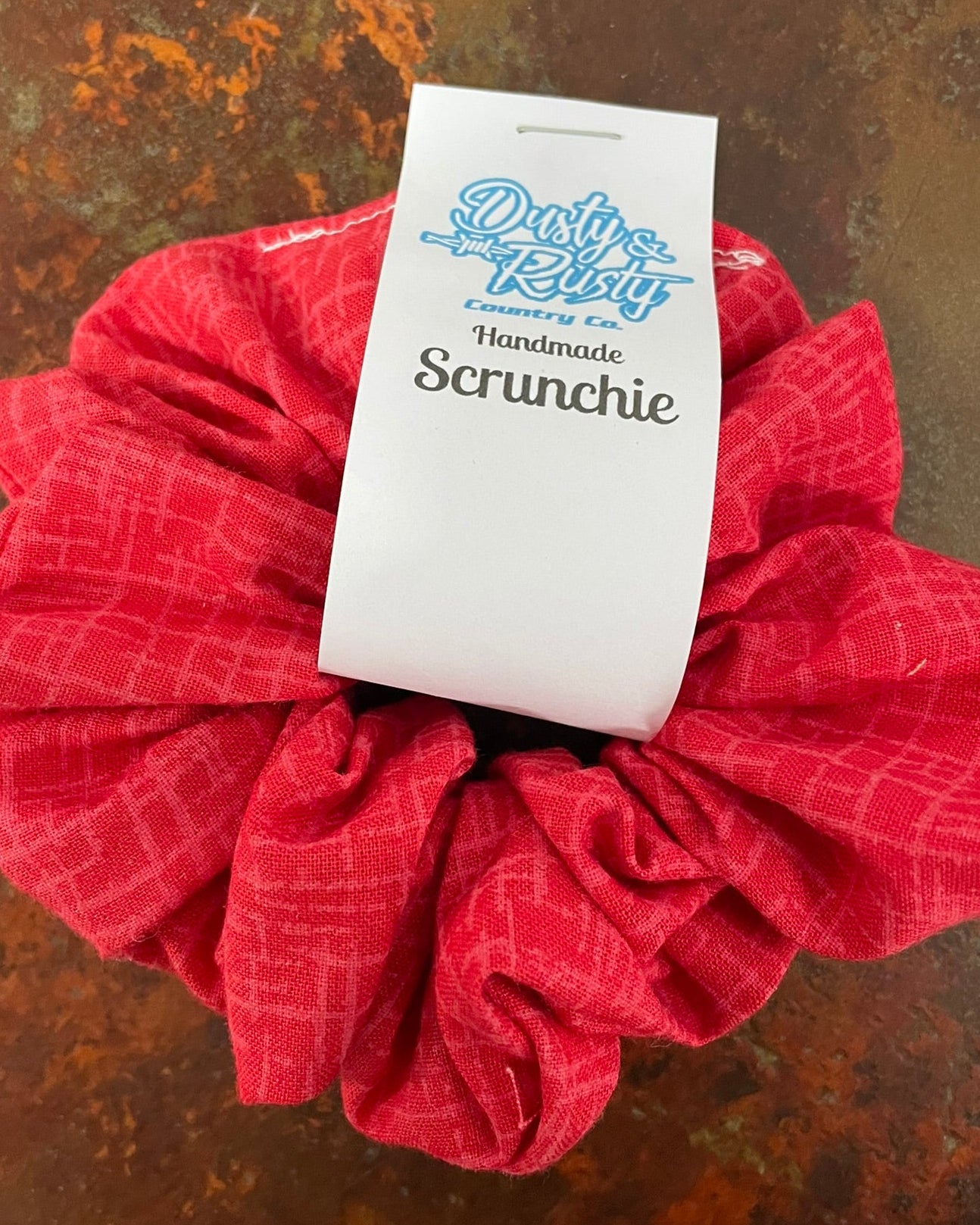 A red fabric scrunchie with a white paper tag that reads 'Dusty Resty Handmade Scrunchie' on a textured brown background.