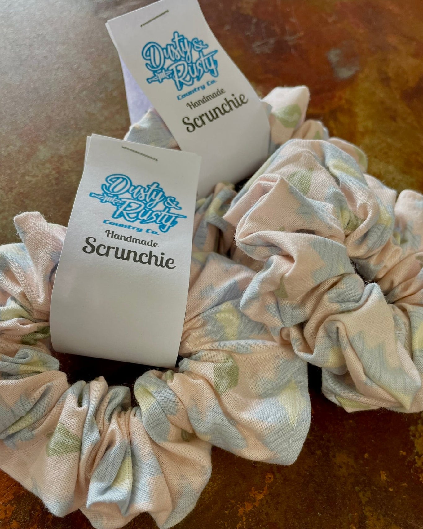 Two fabric scrunchies with white paper tags reading 'Dusty Bunny Handmade Scrunchie' placed on a textured brown surface.