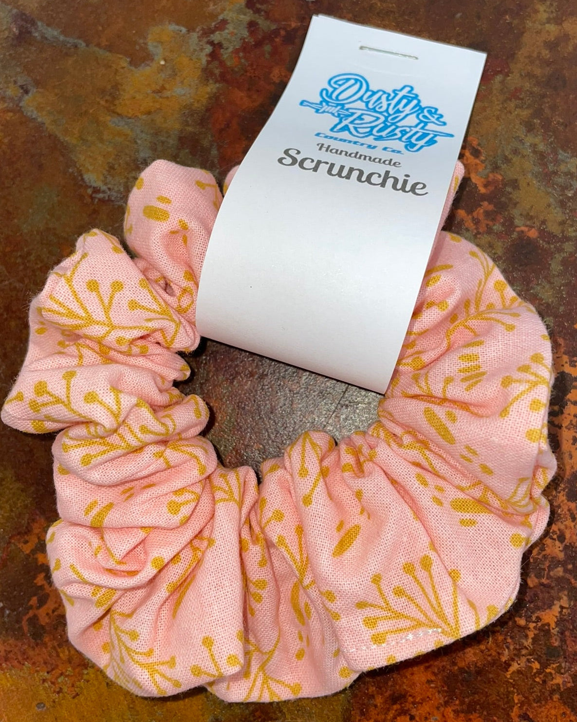 A light pink scrunchie with a yellow botanical pattern, wrapped in a white paper band labeled 'Dusty's Handmade Scrunchie', placed on a textured, rusted metal surface.