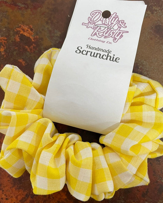 A yellow and white gingham fabric scrunchie with a white paper hang tag reading 'Patty's Rustic Clothing Co. Handmade Scrunchie,' placed on a textured brown surface.